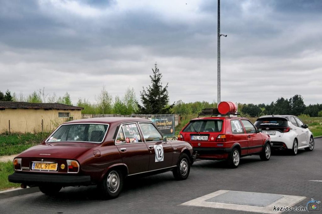 Trzy samochody stoją na poboczu drogi, w tle zielone drzewa i pochmurne niebo.