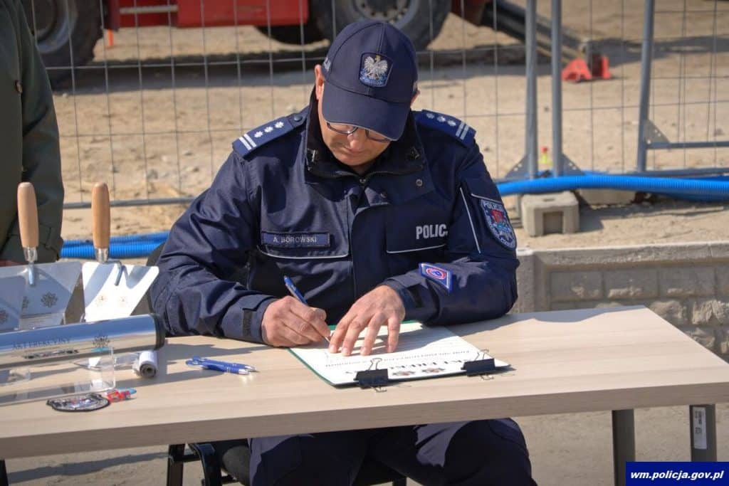Policjant podpisuje dokumenty przy budowie nowoczesnej komendy w Ostr&oacute;dzie, w tle plac budowy.