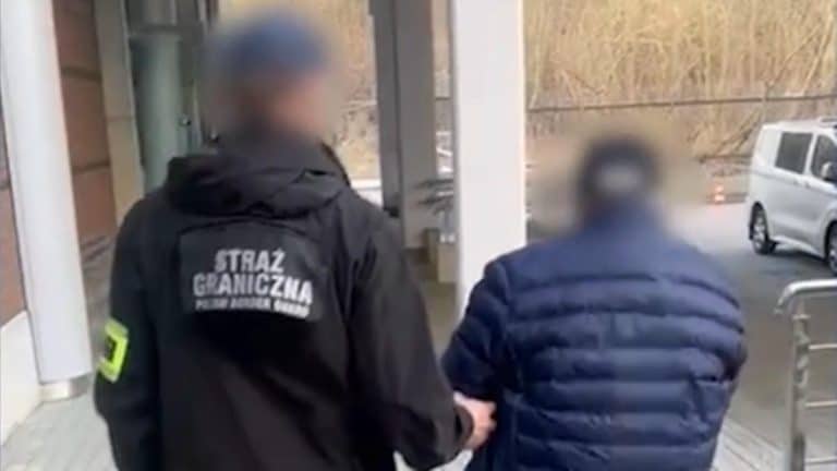 Funkcjonariusz Straży Granicznej prowadzi zatrzymanego mężczyznę, w tle widoczne samochody i wejście do budynku.