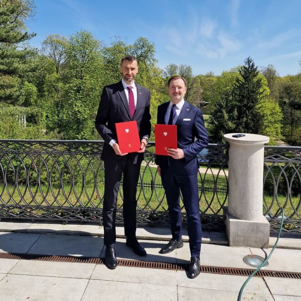 Dw&oacute;ch mężczyzn w garniturach trzyma dokumenty, stojąc na zewnątrz na tle zielonych drzew i nieba.