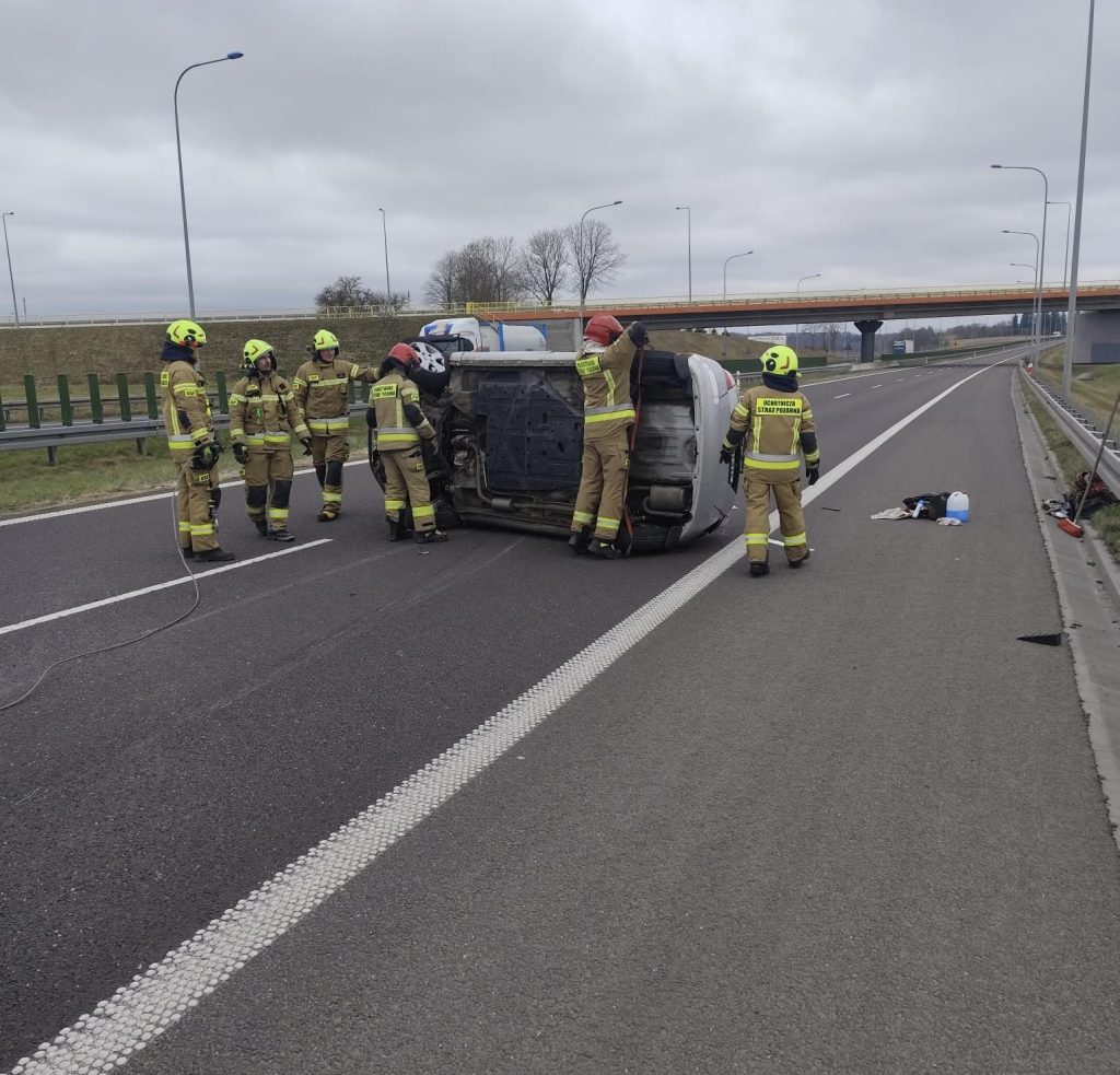 Strażacy wok&oacute;ł przewr&oacute;conego samochodu na drodze S7, w tle most i pochmurne niebo.