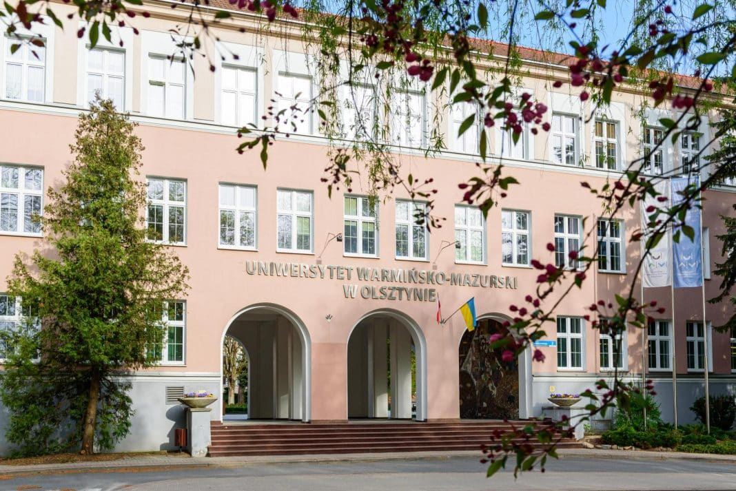 Widok na budynek Wydziału Lekarskiego Uniwersytetu Warmińsko-Mazurskiego w Olsztynie, z drzewami i kwiatami na pierwszym planie.