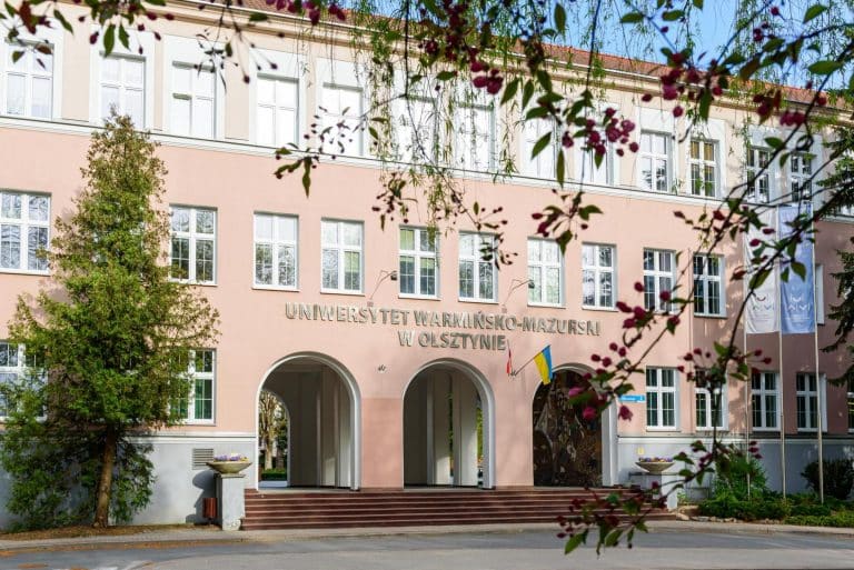 Widok na budynek Wydziału Lekarskiego Uniwersytetu Warmińsko-Mazurskiego w Olsztynie, z drzewami i kwiatami na pierwszym planie.