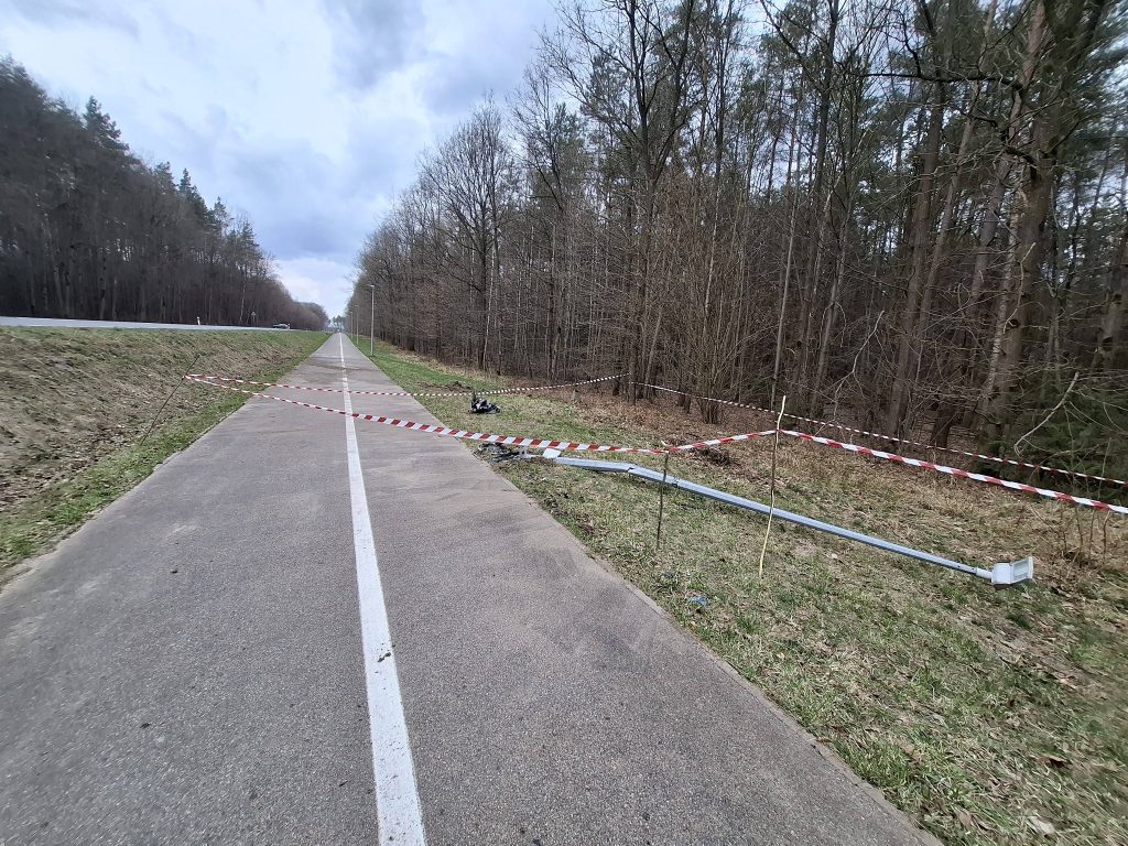 Zabezpieczony odcinek trasy z taśmami i uszkodzonym słupkiem, w tle las i droga.