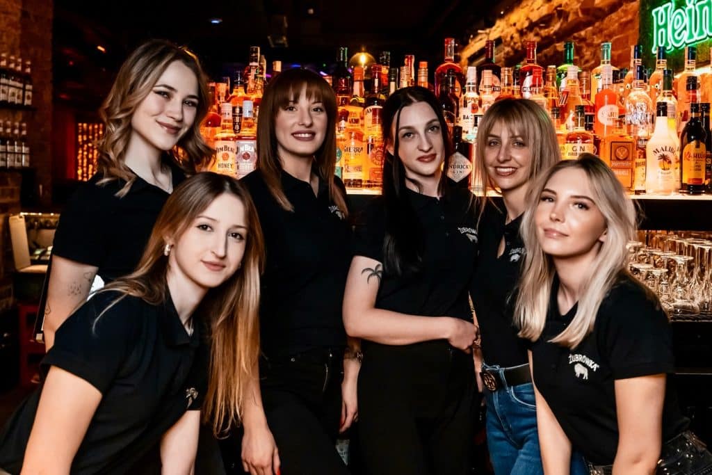 Grupa kobiet w klubie przy barze z szerokim wyborem alkoholi, oświetlenie w klimacie nocnego lokalu.