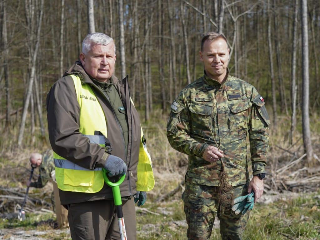 Dwa mężczyźni w mundurach i odblaskowych kamizelkach sadzą drzewa w lesie na Mazurach.