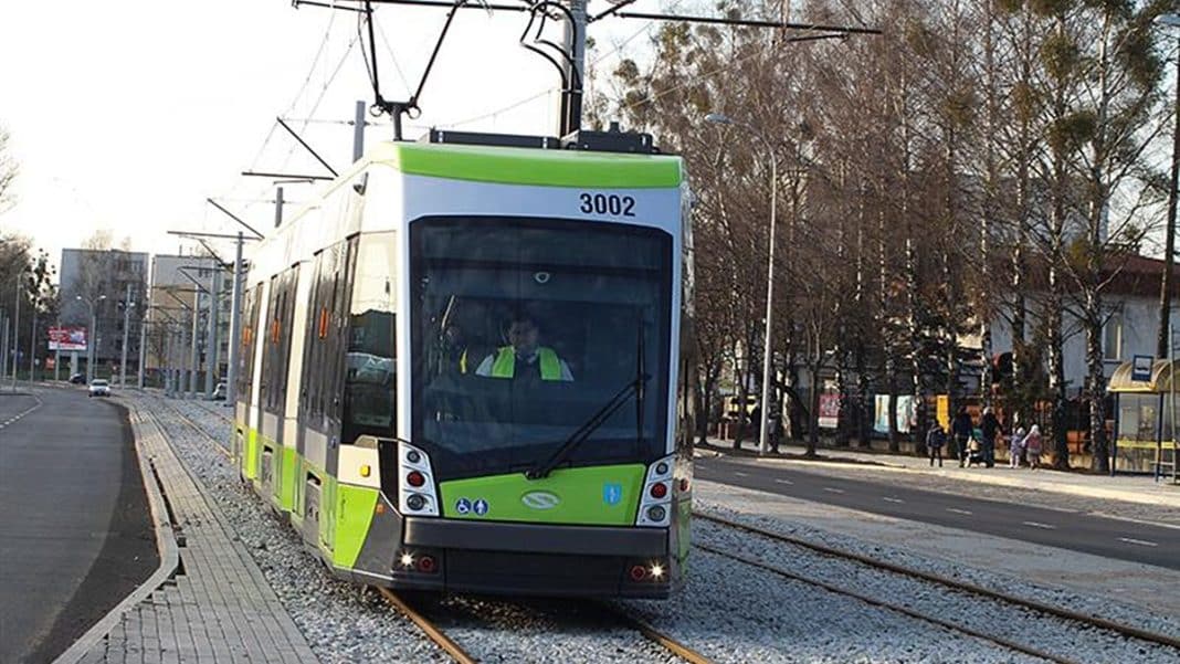 Trolejbus na torach w mieście, w tle drzewa i przystanek autobusowy.