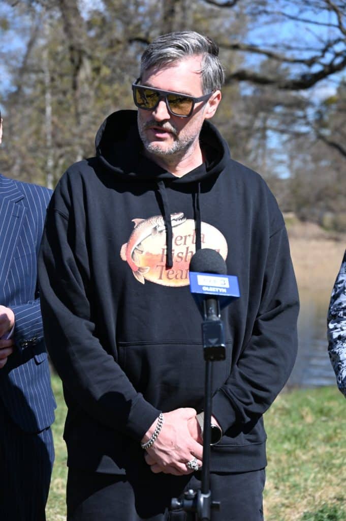 Mężczyzna w czarnej bluzie z logo, okulary przeciwsłoneczne, na tle natury, podczas wydarzenia na świeżym powietrzu.