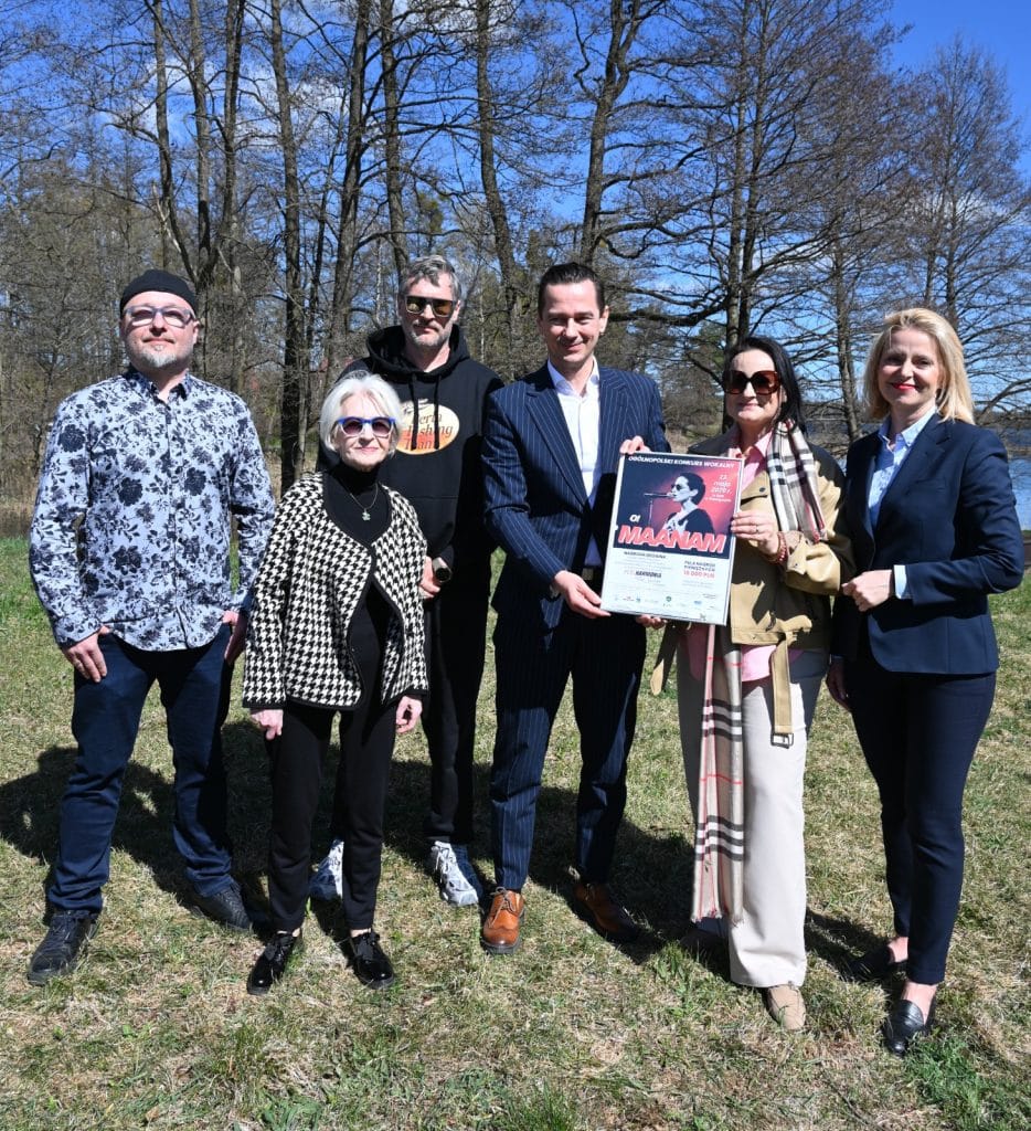Grupa ludzi na zewnątrz, w tle drzewa i niebo, trzymająca plakat związany z konkursem wokalnym.