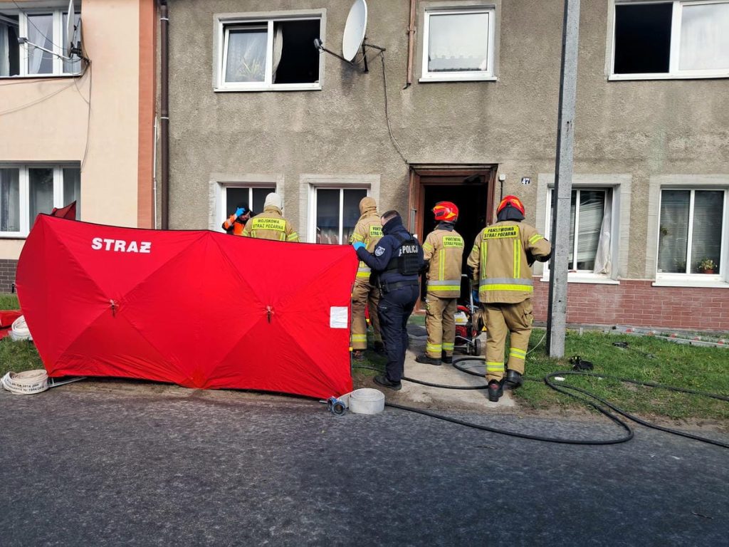 Strażacy wchodzą do mieszkania podczas akcji ratunkowej, na miejscu sprzęt gaśniczy i policja.