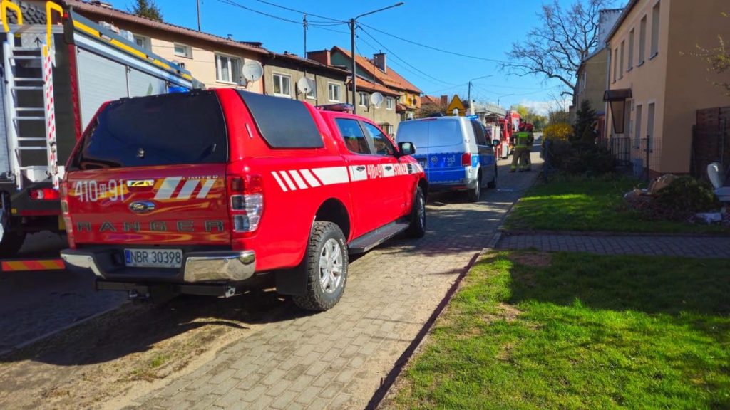 Na ulicy stoją pojazdy straży pożarnej i służb ratunkowych, w tle domy jednorodzinne, słoneczny dzień.