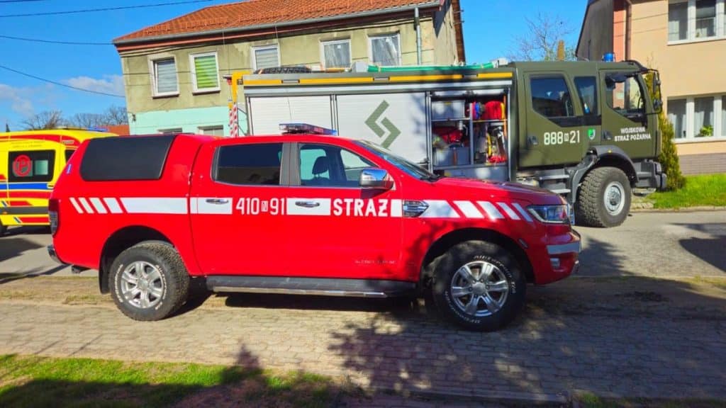 Strażacy wchodzą do mieszkania podczas akcji ratunkowej, obok pojazdy ratunkowe na ulicy.