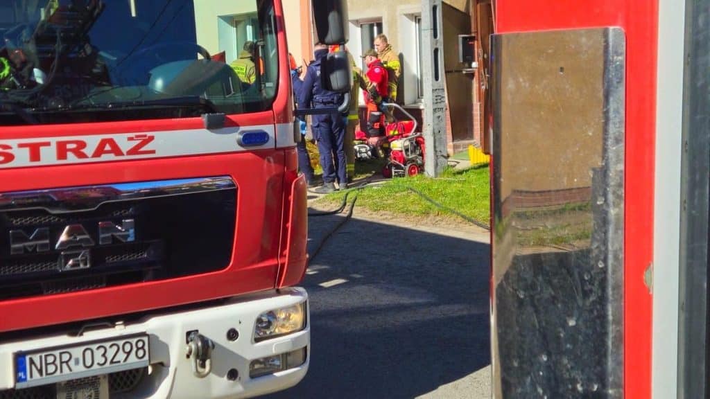 Strażacy w akcji przed wejściem do mieszkania, w tle widać samoch&oacute;d ratunkowy i ekipę ratunkową.