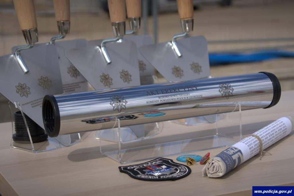 Na zdjęciu widoczny metalowy tubus z napisem o budowie nowej komendy policji w Ostr&oacute;dzie, obok odznaki i dokumenty.