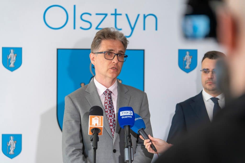 Mężczyzna w garniturze przemawia podczas konferencji w Olsztynie, za nim herb miasta i napis "Olsztyn".
