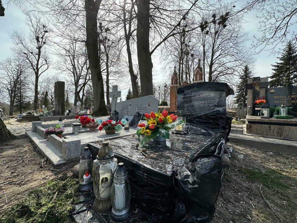 Nagrobek owinięty folią na cmentarzu, wok&oacute;ł kwiaty i znicze, w tle drzewa i inne nagrobki.