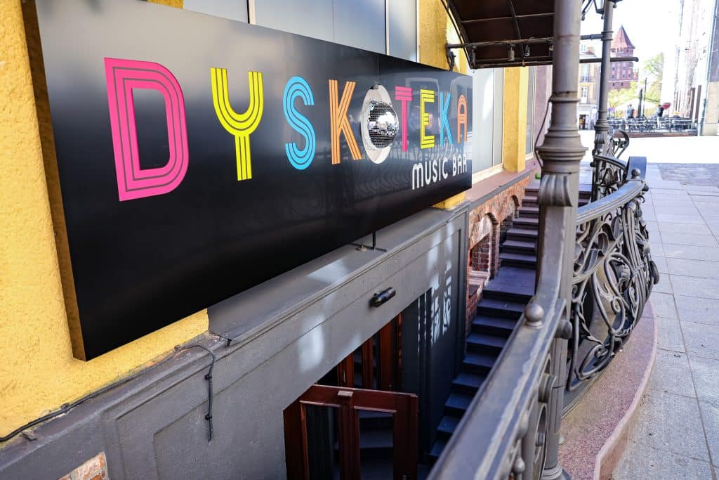 Widok na wejście do nowego klubu Dyskoteka Music Bar na star&oacute;wce w Olsztynie, z kolorowym logo na ścianie.