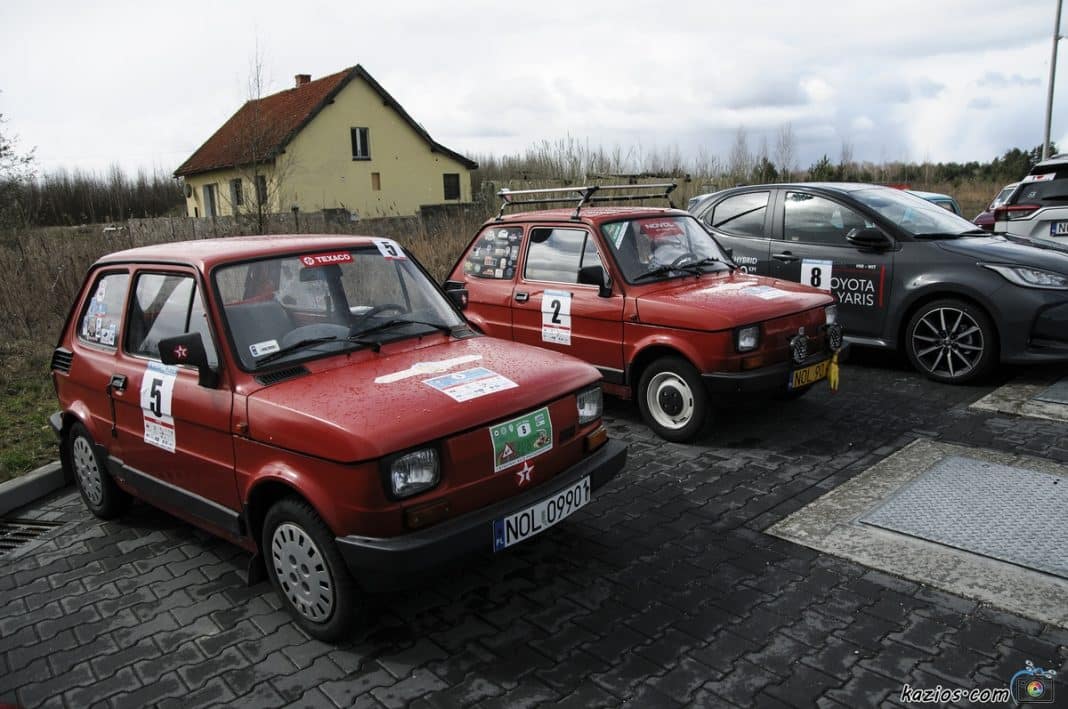 Dwa stare samochody na parkingu, obok nowoczesny pojazd, w tle dom i chmury na niebie.