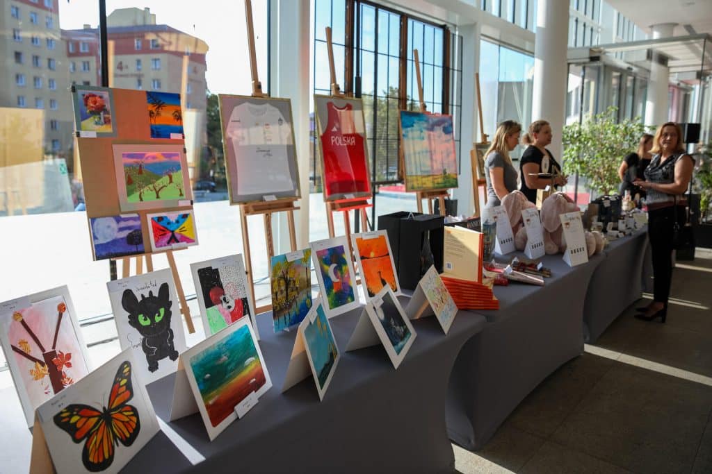 Wystawa obraz&oacute;w i rękodzieła podczas wydarzenia w Olsztynie, z widokiem na okna i ludzi oglądających prace.