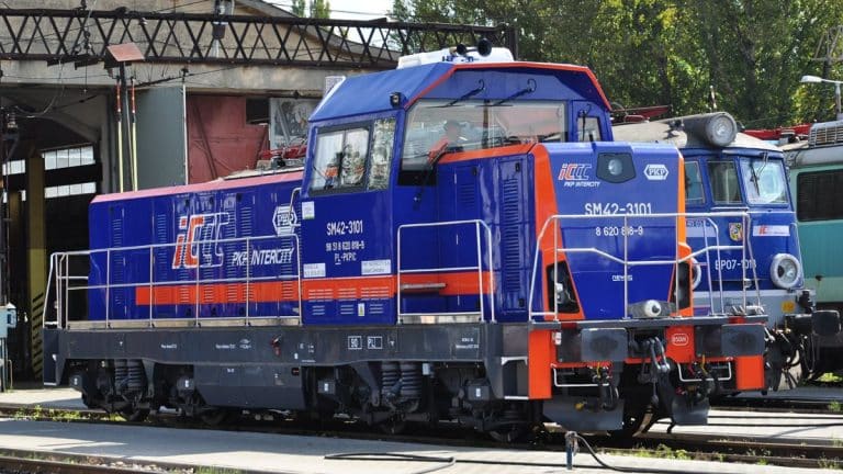 Lokomotywa kolejowa na torze w zajezdni, w tle inne pojazdy i zadaszenie.