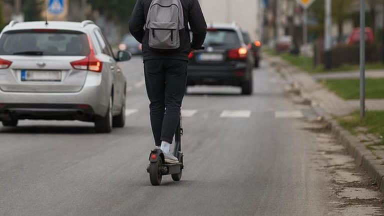 Młody mężczyzna na hulajnodze elektrycznej jedzie środkiem jezdni, za nim samochody, w mieście.