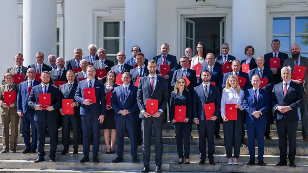 Grupa os&oacute;b w formalnych strojach na schodach przed budynkiem, niekt&oacute;re trzymają czerwone foldery.