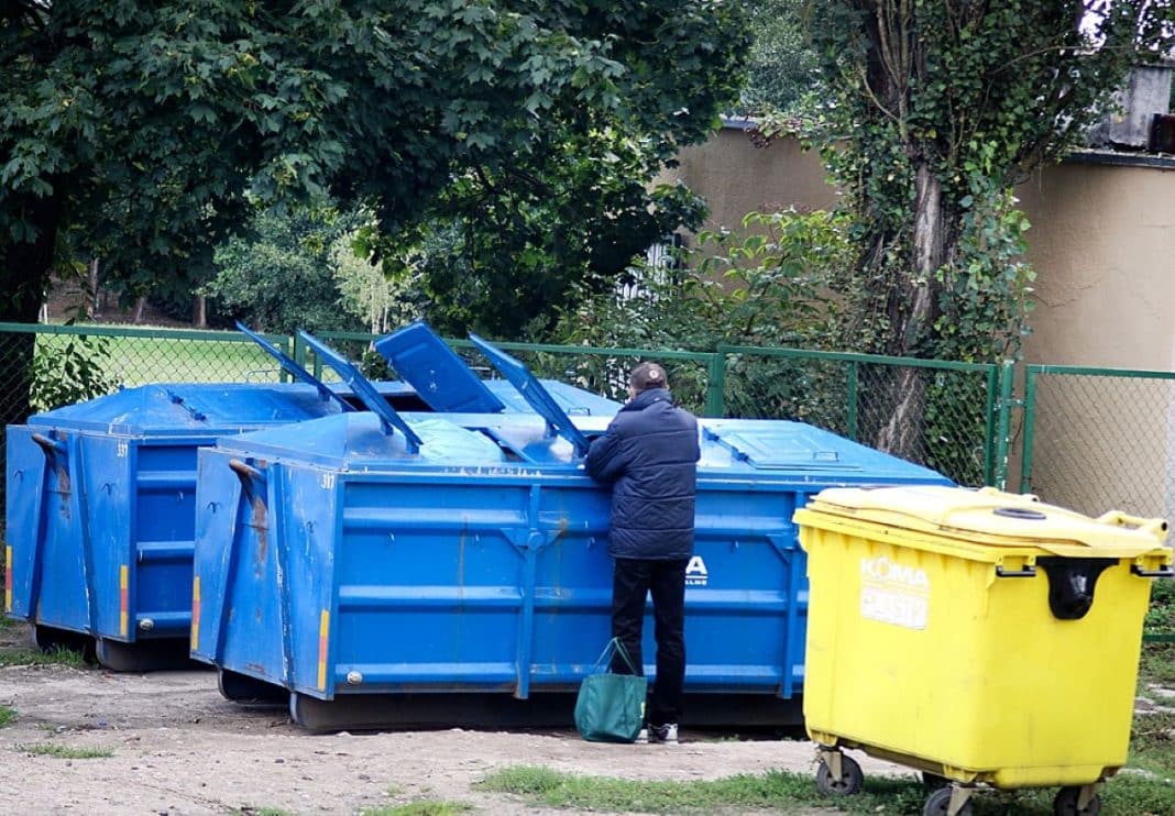Mężczyzna stoi obok kontener&oacute;w na śmieci przy zielonej siatce i drzewach w tle.