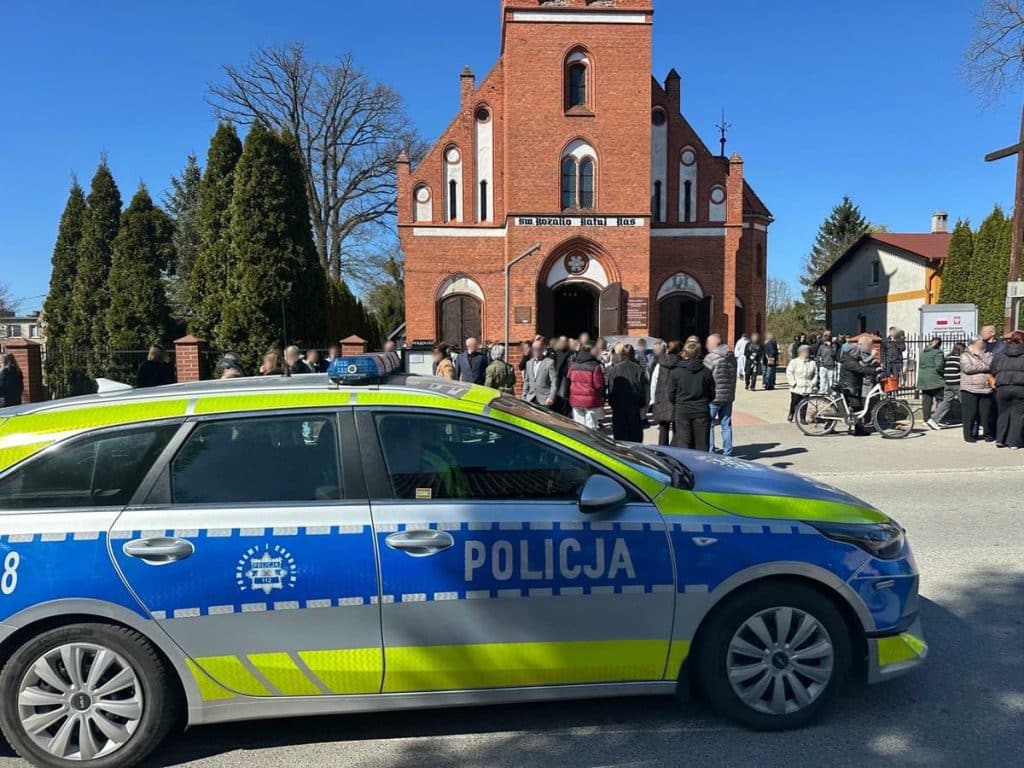 Tłum ludzi przed kościołem podczas pogrzebu 14-letniego chłopca, na pierwszym planie radiow&oacute;z policji.