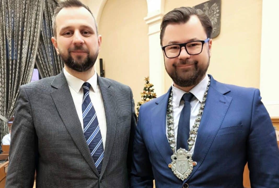 Dw&oacute;ch mężczyzn w garniturach na oficjalnym spotkaniu, jeden z nich z medalem na szyi, w tle dekoracje i herb miasta.