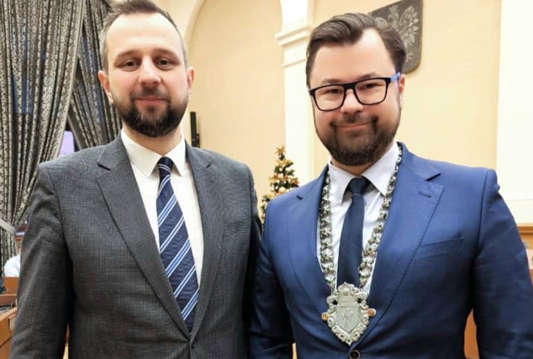 Dw&oacute;ch mężczyzn w garniturach na oficjalnym spotkaniu, jeden z nich z medalem na szyi, w tle dekoracje i herb miasta.