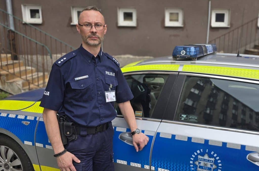 Policjant stojący obok radiowozu na tle budynku, w mundurze, na ulicy.