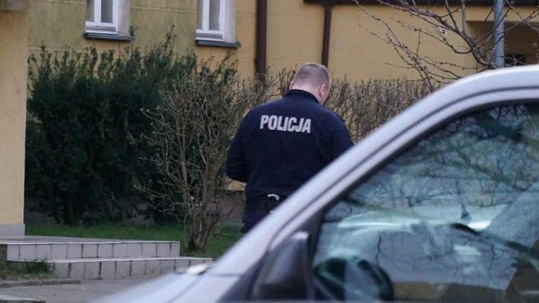 Policjant na miejscu zdarzenia w pobliżu mieszkania, w tle drzewa i budynek.