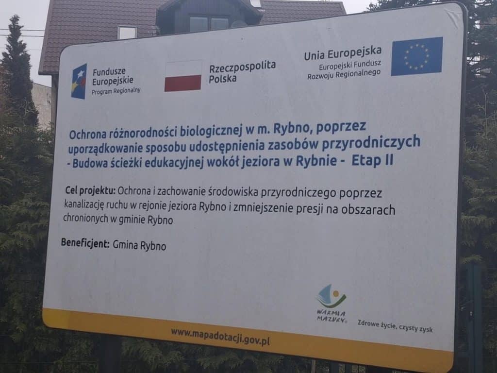 Tablica informacyjna o projekcie ochrony przyrody wok&oacute;ł jeziora Rybno, z logo funduszy europejskich.