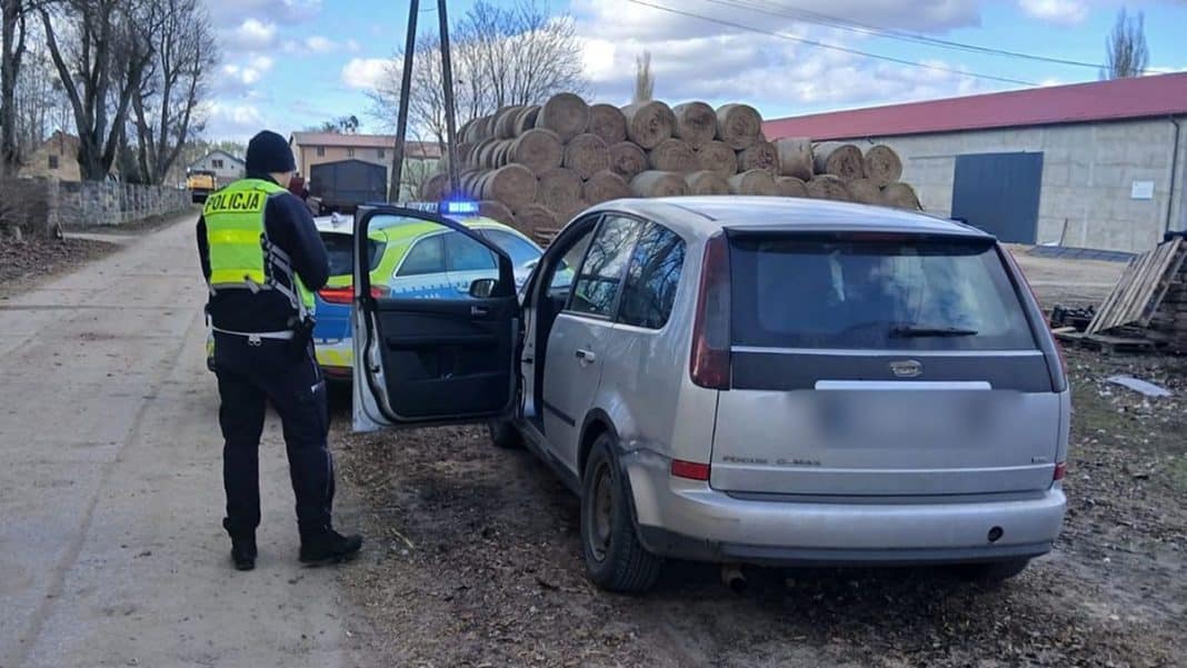 Policjant zatrzymuje samoch&oacute;d na wiejskiej drodze, w tle stos bel siana i budynki.