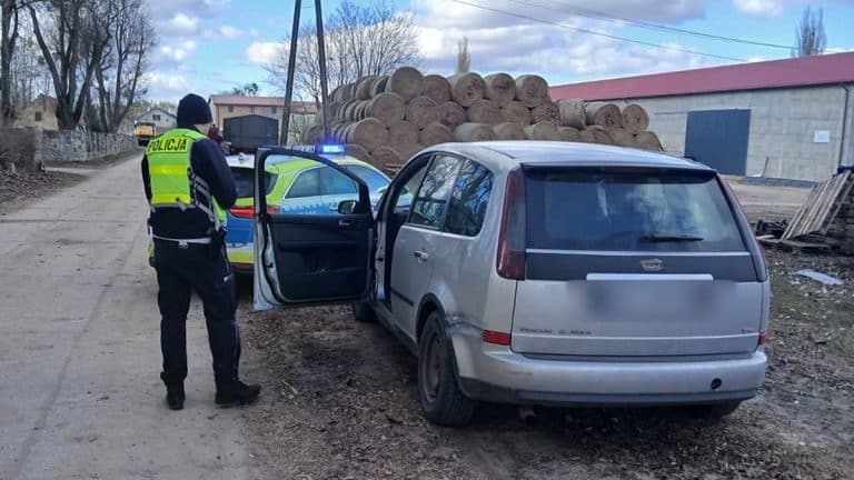 Policjant zatrzymuje samoch&oacute;d na wiejskiej drodze, w tle stos bel siana i budynki.
