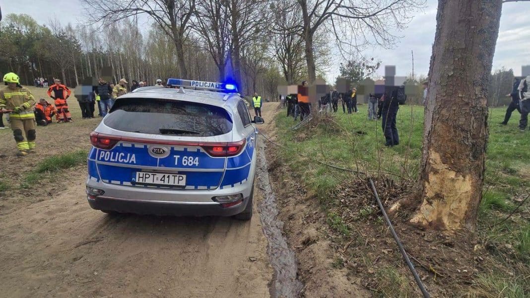 Rozbity samoch&oacute;d po wypadku na odcinku rajdu, otoczony przez ratownik&oacute;w i kibic&oacute;w, w pobliżu drzewa na szutrowej trasie.