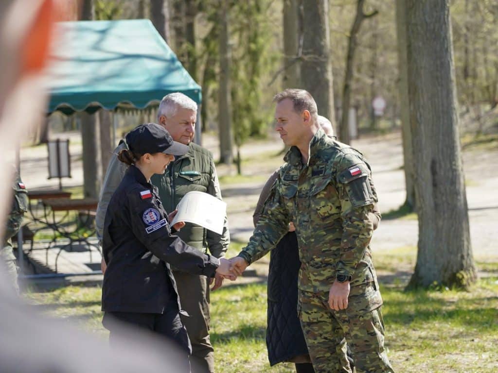 Uczestnicy akcji sadzenia drzew w lesie, w tym strażnik i funkcjonariusz, podczas wydarzenia na Mazurach.