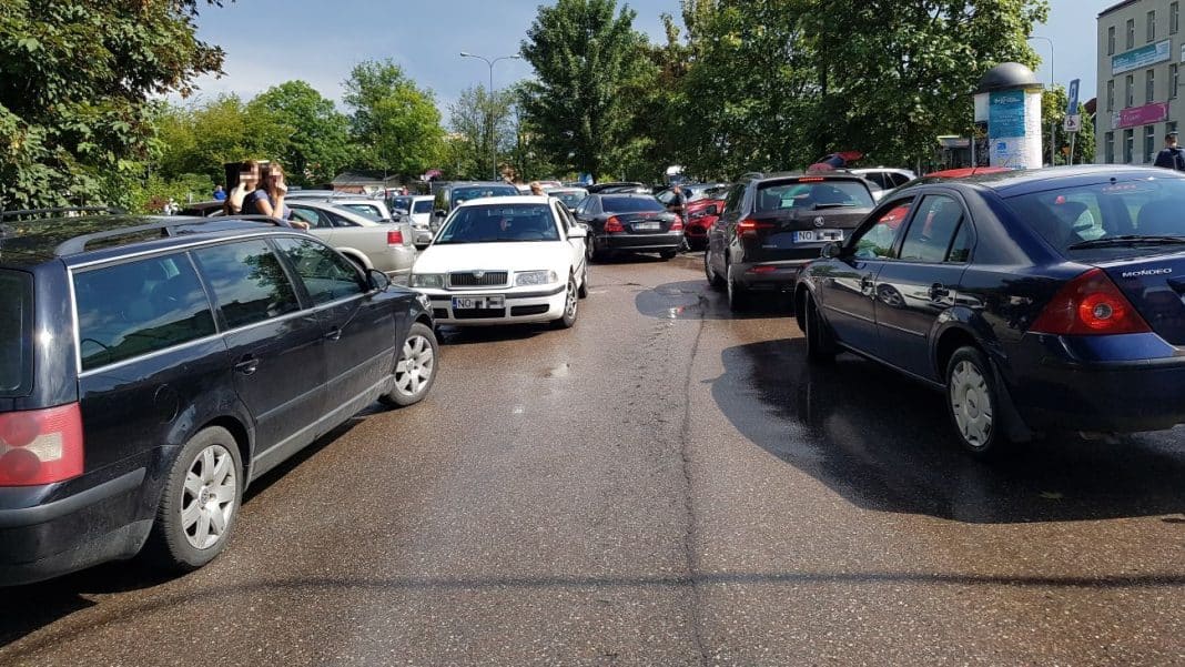 Zabłocona i mokra ulica z zaparkowanymi samochodami, na tle zielonych drzew i budynk&oacute;w.