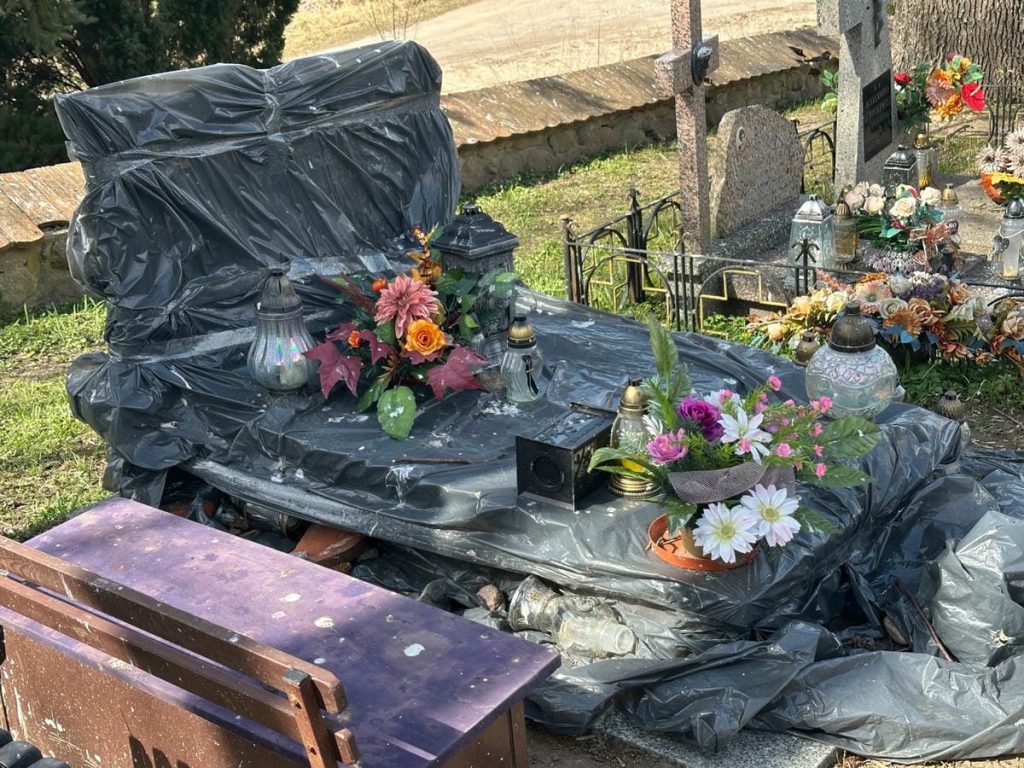 Nagrobek przykryty czarnym folią, ozdobiony kwiatami i lampionami, na tle innych grob&oacute;w na cmentarzu.