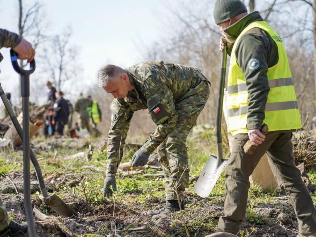 Uczestnicy sadzą drzewa podczas akcji w Wilczym Szańcu na Mazurach, w tle las i grupa ludzi.