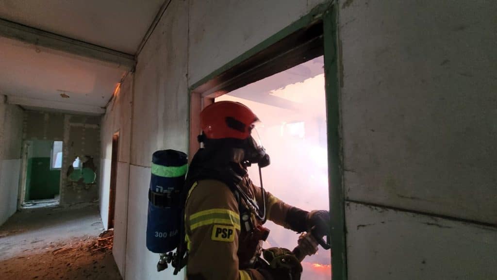 Strażak w hełmie i ubraniu ochronnym walczy z pożarem w pustostanie, widok przez okno na płomienie.