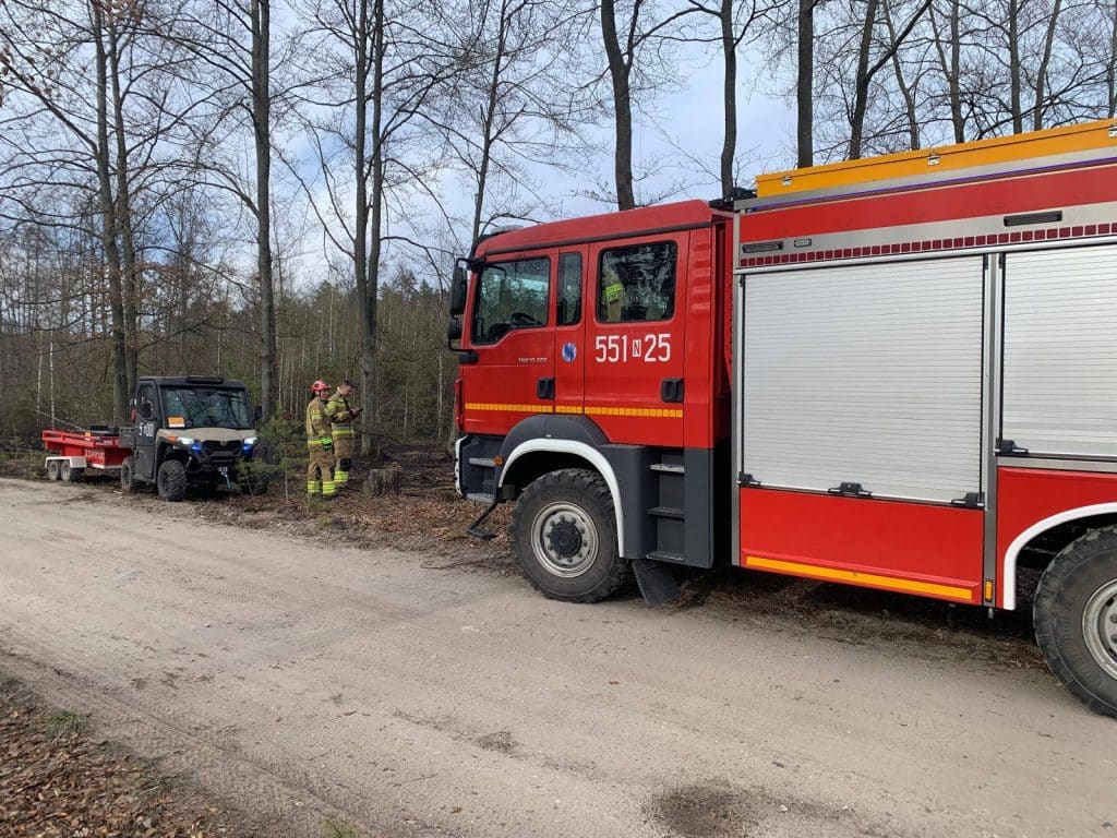 Strażacy gaszą pożar lasu w okolicach Rucianego-Nidy, w tle drzewa i pojazdy ratunkowe.