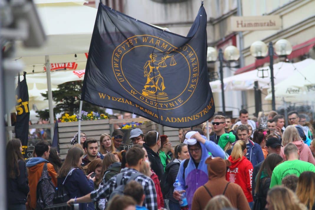 Grupa student&oacute;w na ulicy z flagą uniwersytetu w tle, tłum ludzi na wydarzeniu.