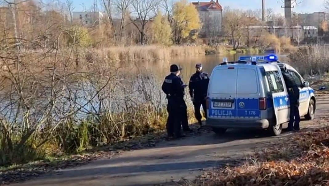 Policjanci stoją przy radiowozie nad Jeziorem Długim w Olsztynie, w tle drzewa i budynki.