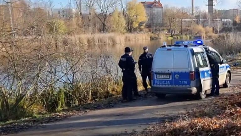 Policjanci stoją przy radiowozie nad Jeziorem Długim w Olsztynie, w tle drzewa i budynki.