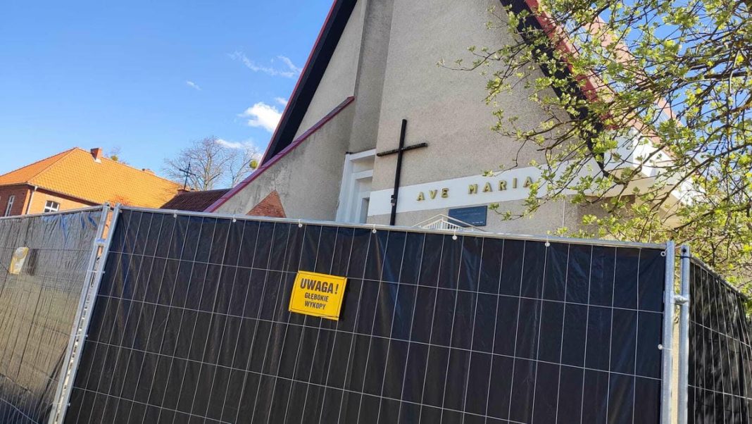 Teren wok&oacute;ł kościoła otoczony ogrodzeniem, widoczna tablica ostrzegawcza o wykopach.