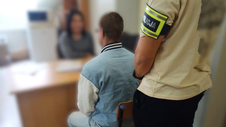 Policjant rozmawia z zatrzymanym mężczyzną w pomieszczeniu, w tle kobieta przy biurku.