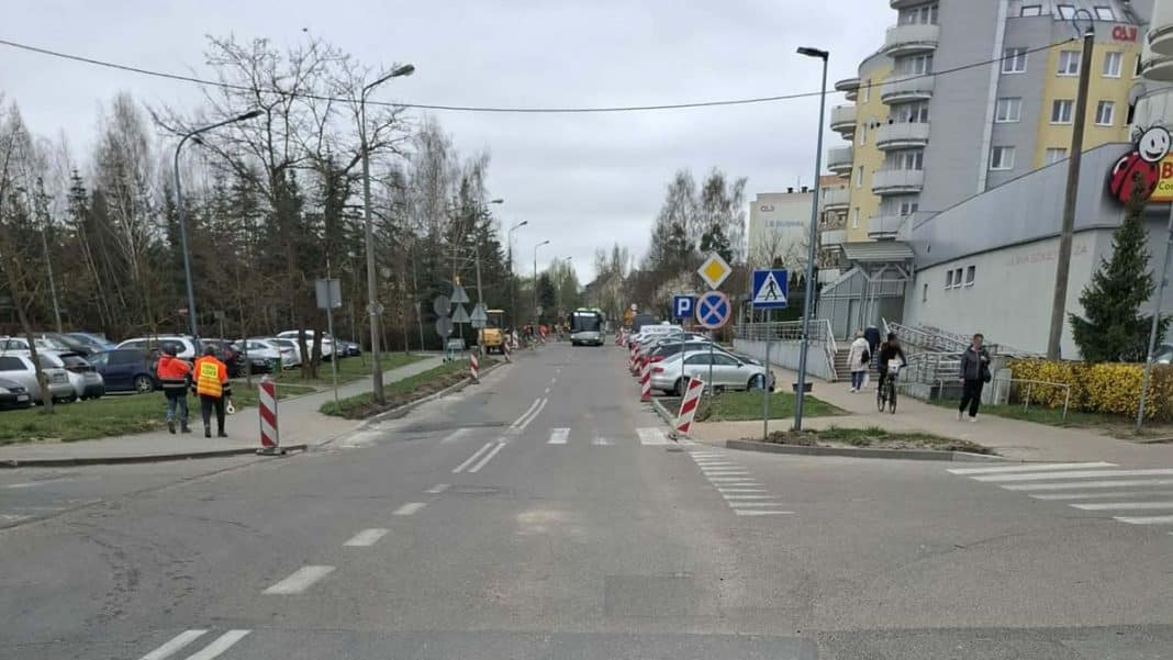 Ulica Iwaszkiewicza w Olsztynie z oznaczeniami i barierkami, w tle pojazdy i budynki mieszkalne.