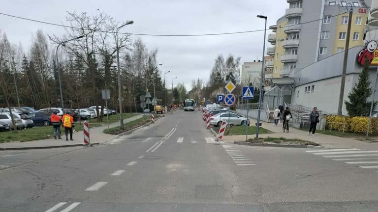 Ulica Iwaszkiewicza w Olsztynie z oznaczeniami i barierkami, w tle pojazdy i budynki mieszkalne.