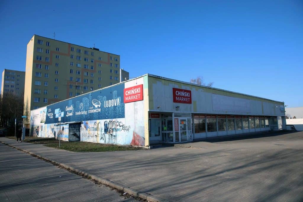 Budynek handlowy przy Hali Urania w Olsztynie, na tle wysokich blok&oacute;w mieszkalnych, z graffiti na ścianie.